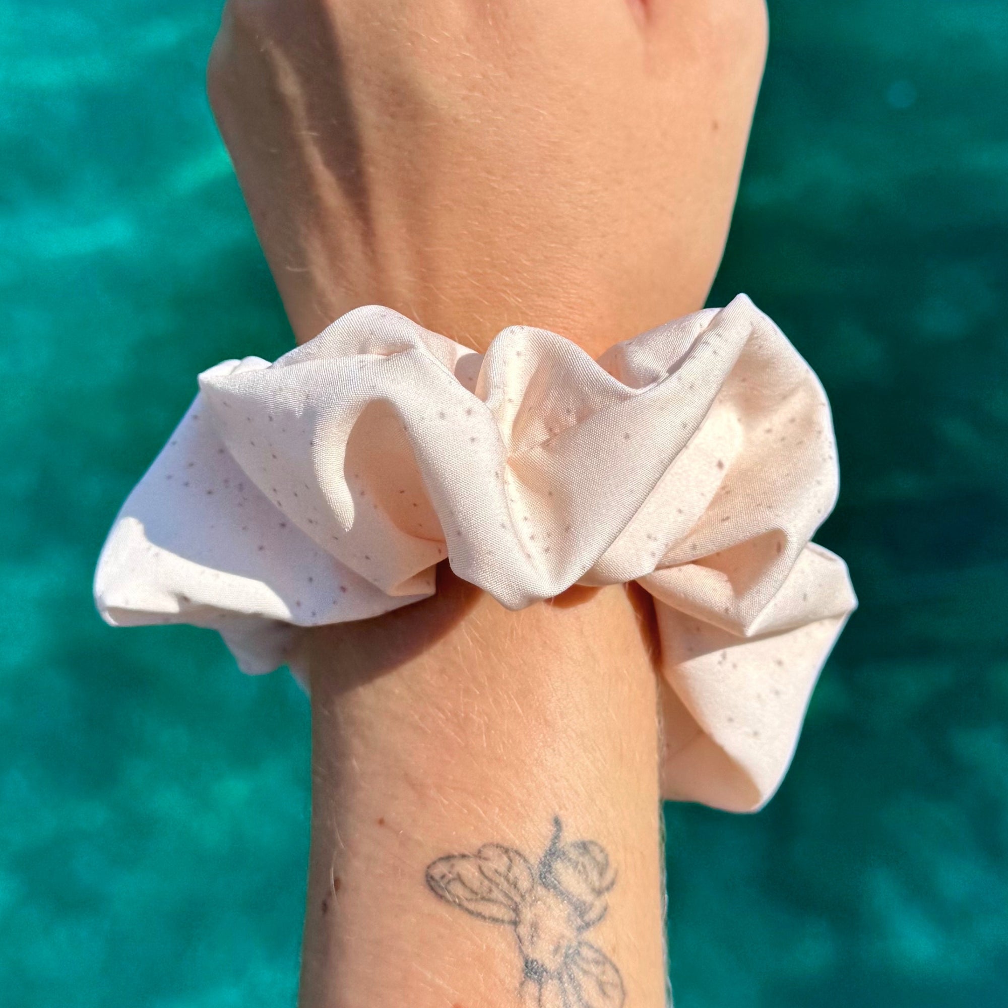 A close-up of a recycled fabric scrunchie worn on a person's wrist, with a turquoise water background and a small tattoo visible on the forearm.