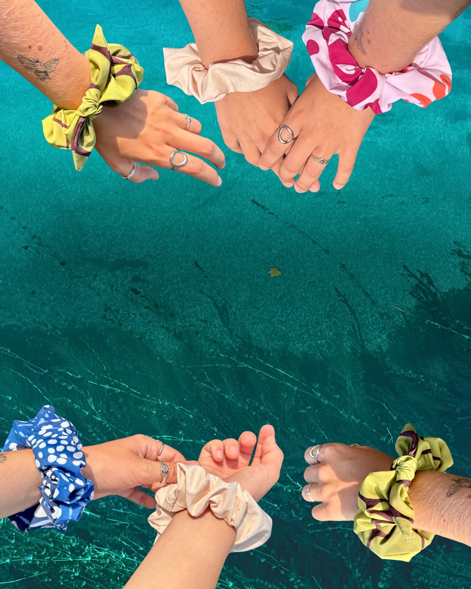 Four hands wearing recycled Kelpia scrunchies above clear turquoise sea water