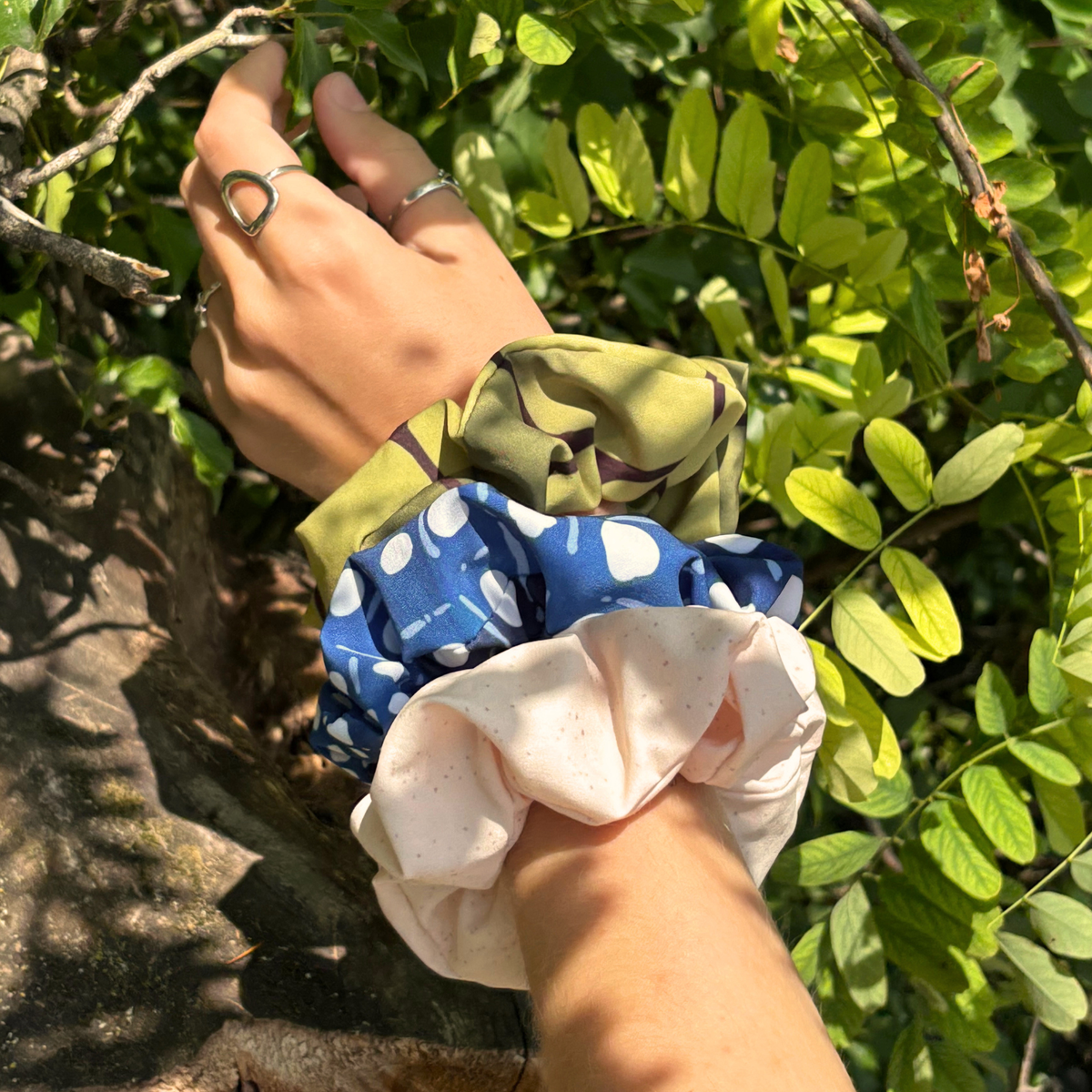 Three recycled fabric hair scrunchies in different prints (olive green camo, blue with white spots, and light pink) worn on a person's wrist and hand, shown outdoors among green leaves.