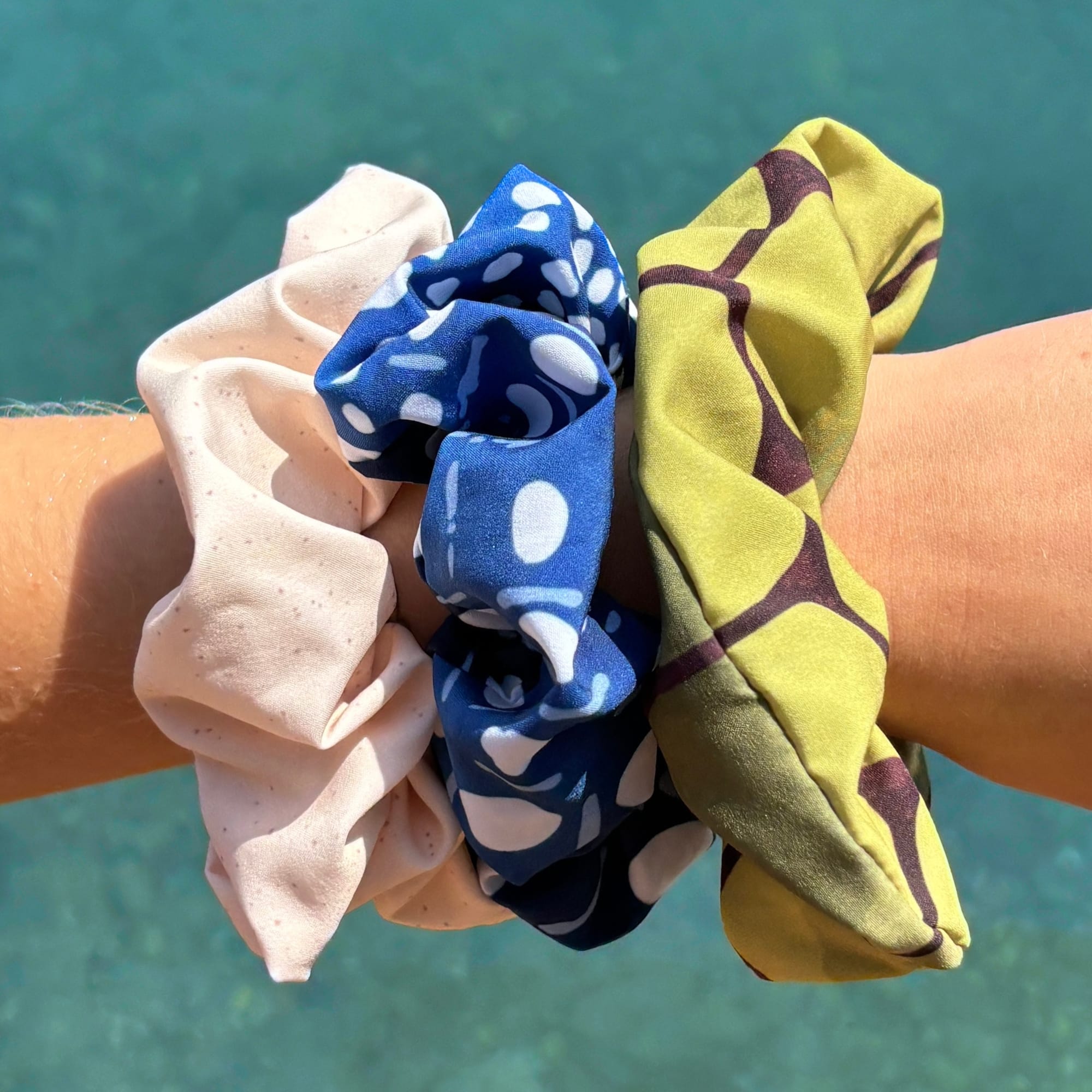 Three fabric hair scrunchies worn on a person's wrist against a turquoise water background. The scrunchies are made of different patterned fabrics: one is light beige with tiny speckles, one is royal blue with white abstract shapes, and one is olive green with dark brown abstract lines.