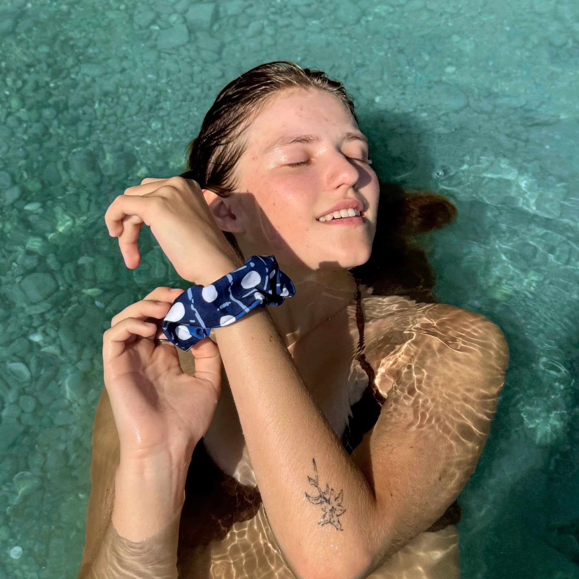 A woman floating in clear turquoise water wears a navy blue scrunchie with white polka dots and a white abstract line pattern.