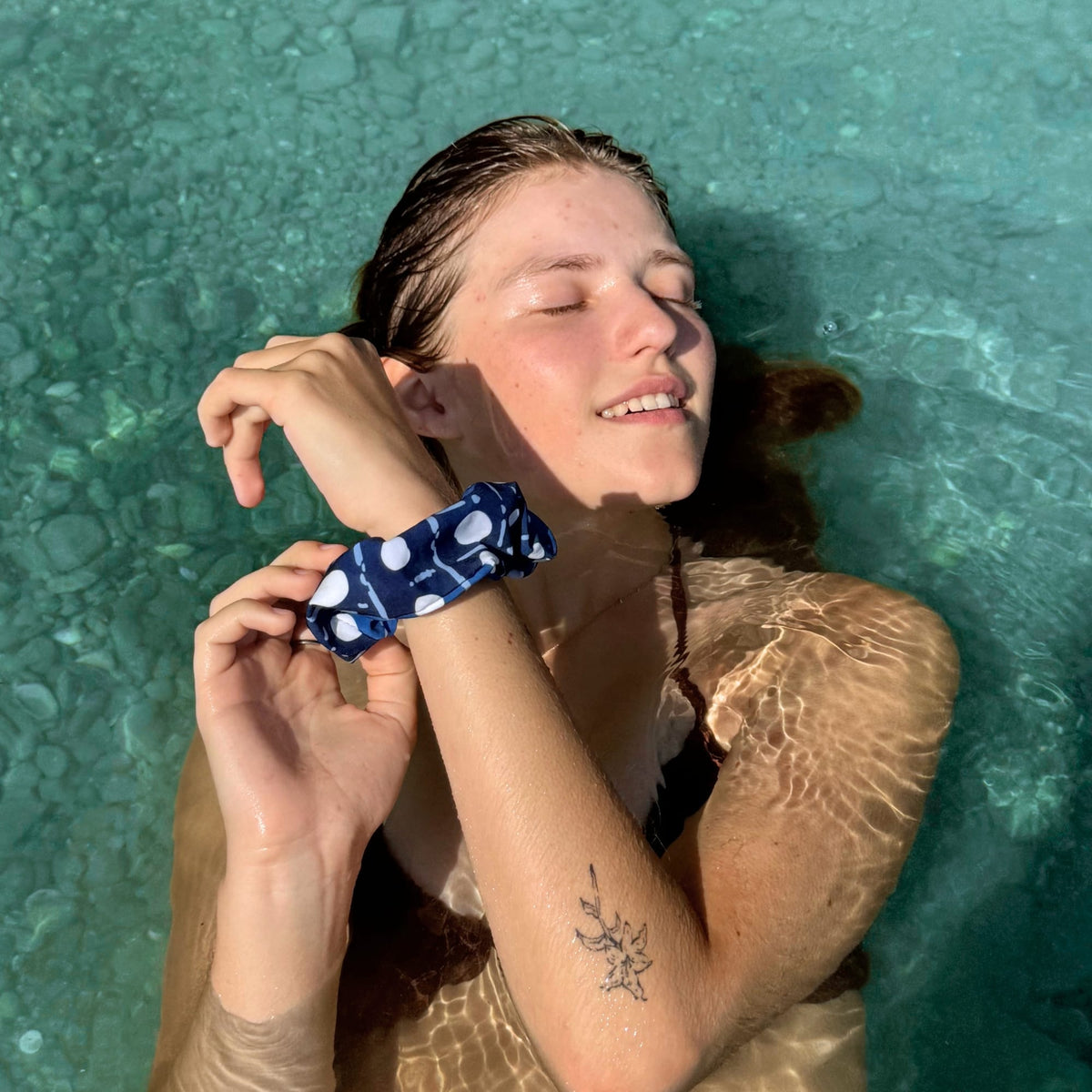 A woman floating in clear turquoise water wears a navy blue scrunchie with white polka dots and a white abstract line pattern.