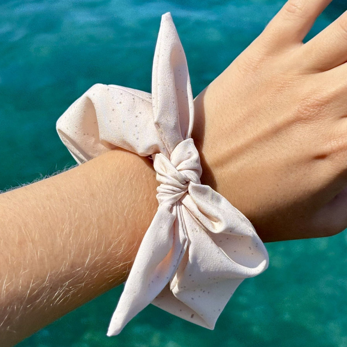 A light pink fabric scrunchie with tiny dark speckles tied around a person's wrist, with a turquoise ocean background. Removable bow