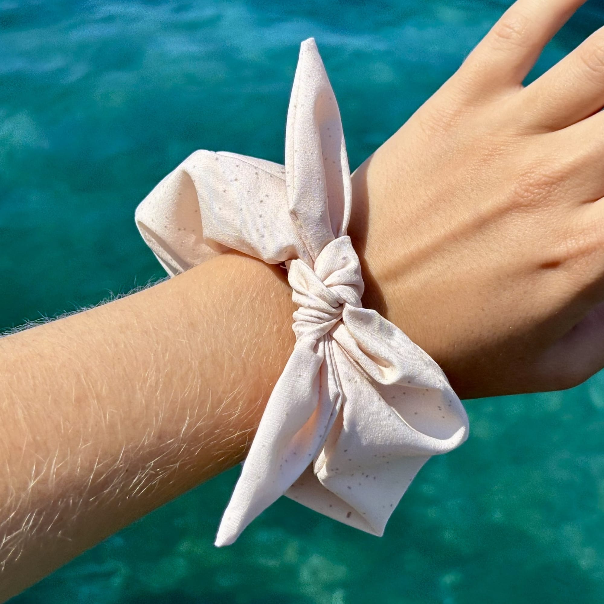 A light sandy fabric scrunchie with tiny dark speckles tied around a person's wrist, with a turquoise ocean background. Made of recycled fabric