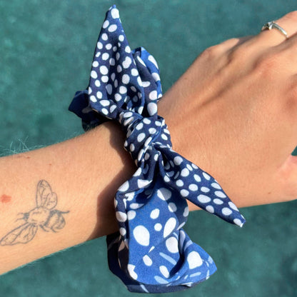 A blue and white polka dot fabric scrunchie Whale SHark design worn on a person's wrist, with a tattoo visible on the forearm and a ring on the hand, set against a teal water background.