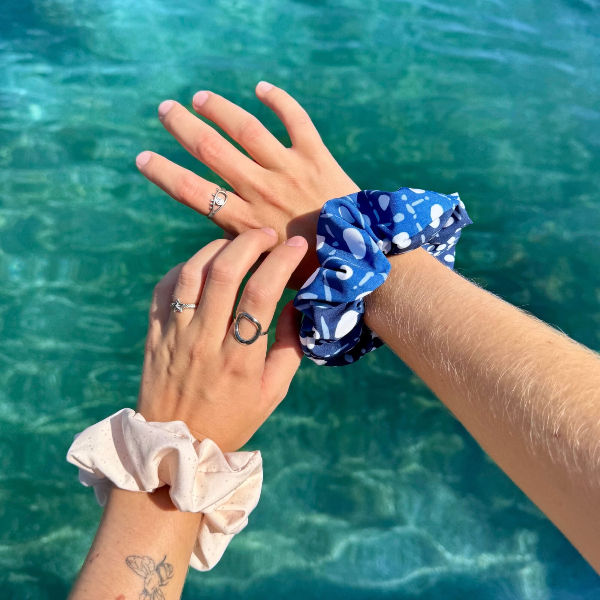 Two recycled fabric scrunchies in different colors and patterns are shown on wrists against a tropical turquoise ocean background. One scrunchie is light pink, one is blue with white abstract spots, and one is blue with white wavy lines.